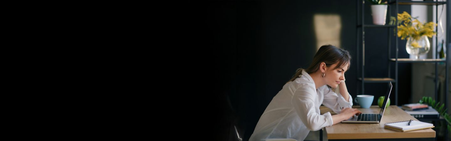 Mlada žena sjedi za stolom i koncentrirano radi na laptopu, s bilježnicom i šalicom pored sebe. Pozadina je tamna, a fokus je na mladoj ženi.<br/>