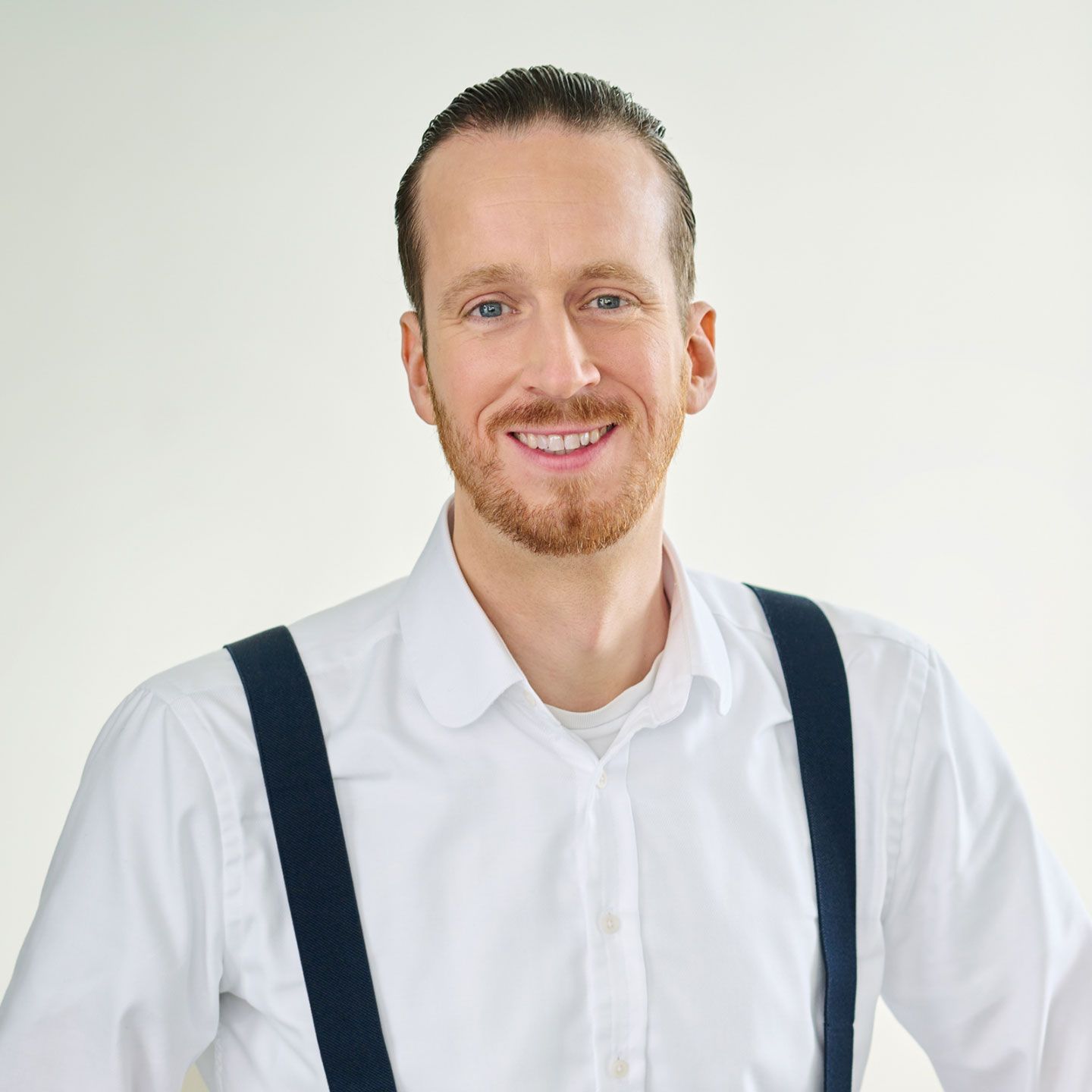 Sebastian Pollmer, muškarac, stoji u bijeloj košulji i plavim tregerima u svijetloj sobi. 