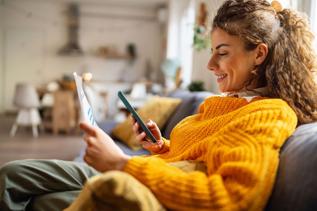 Nasmijana mlada žena s kovrčavom kosom, u udobnom žutom džemperu, sjedi na kauču u svom dnevnom boravku i gleda u svoj pametni telefon dok drži papirnati dokument.
