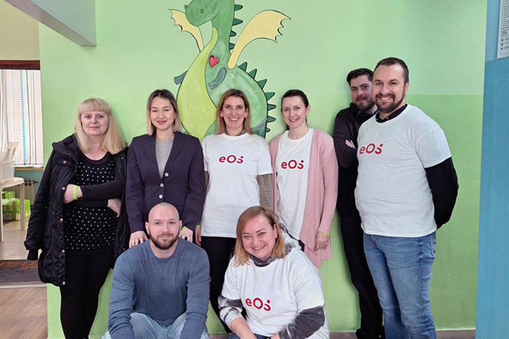 Osmero ljudi stoji ispred zida na kojem je naslikan zeleni zmaj. Ljudi su raspoređeni u dva reda: dva su smještena sprijeda, a šest ih stoji straga. Četiri osobe nose bijele majice s crvenim EOS-ovim logotipom. Grupa se smiješi prema kameri. Na podu, sprijeda s desne strane, vidimo nekoliko boca i kartona od pića.