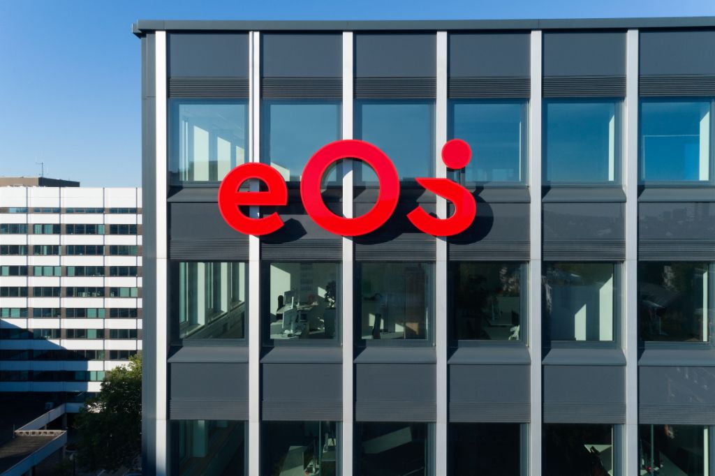 An exterior, mid-angle shot of a contemporary building facade, featuring a prominent red "eos" logo mounted above a series of windows on a dark grey panel. The building has a modern design with vertical white accents separating sections of windows and panels. The sky above is clear and blue. In the background, another multi-story building with numerous windows is partially visible on the left side of the frame, suggesting an urban or corporate environment.