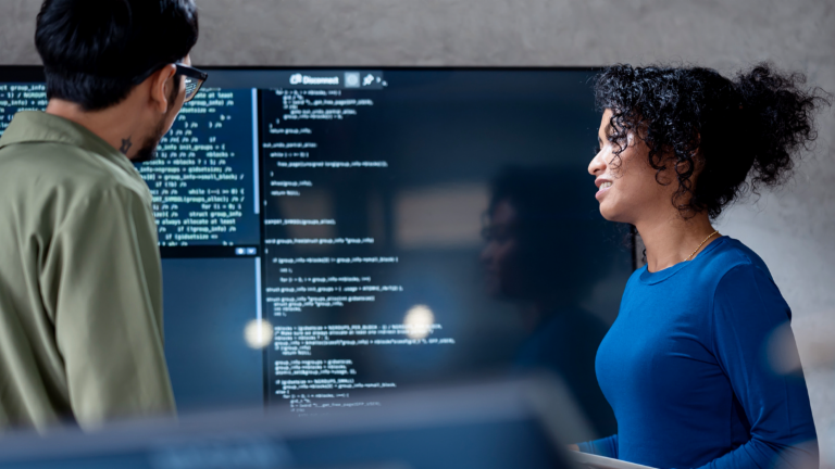 AI u naplati dugova: Na ovoj slici, dva stručnjaka zajednički analiziraju programski kod za automatizovane procese na velikom ekranu. Scena ilustruje praktičnu primenu veštačke inteligencije (AI) i robotizovane automatizacije procesa (RPA) u upravljanju dugovima — primer digitalne transformacije u upravljanju potraživanjima.