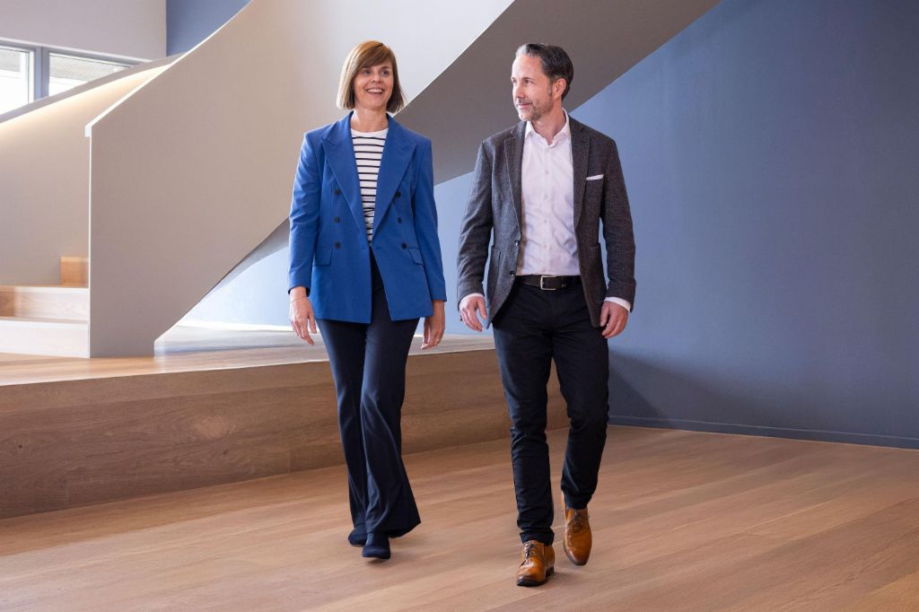 The CEO in a gray blazer and the CFO of the EOS Group in a blue blazer walk through a modern office. The space features large windows, a curved wall, and stylish hanging lamps.