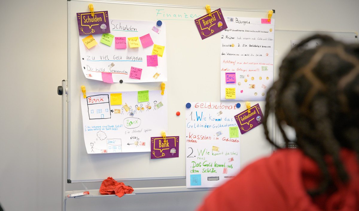 A photo shows post-its and paper notes on a board with terms like “Debt”, “Cash”, “Bank” and “ATM”. Underneath, the text reads: “In the ManoMoneta program developed by the EOS finlit foundation, schoolchildren aged 9 to 13 can learn about financial products.“