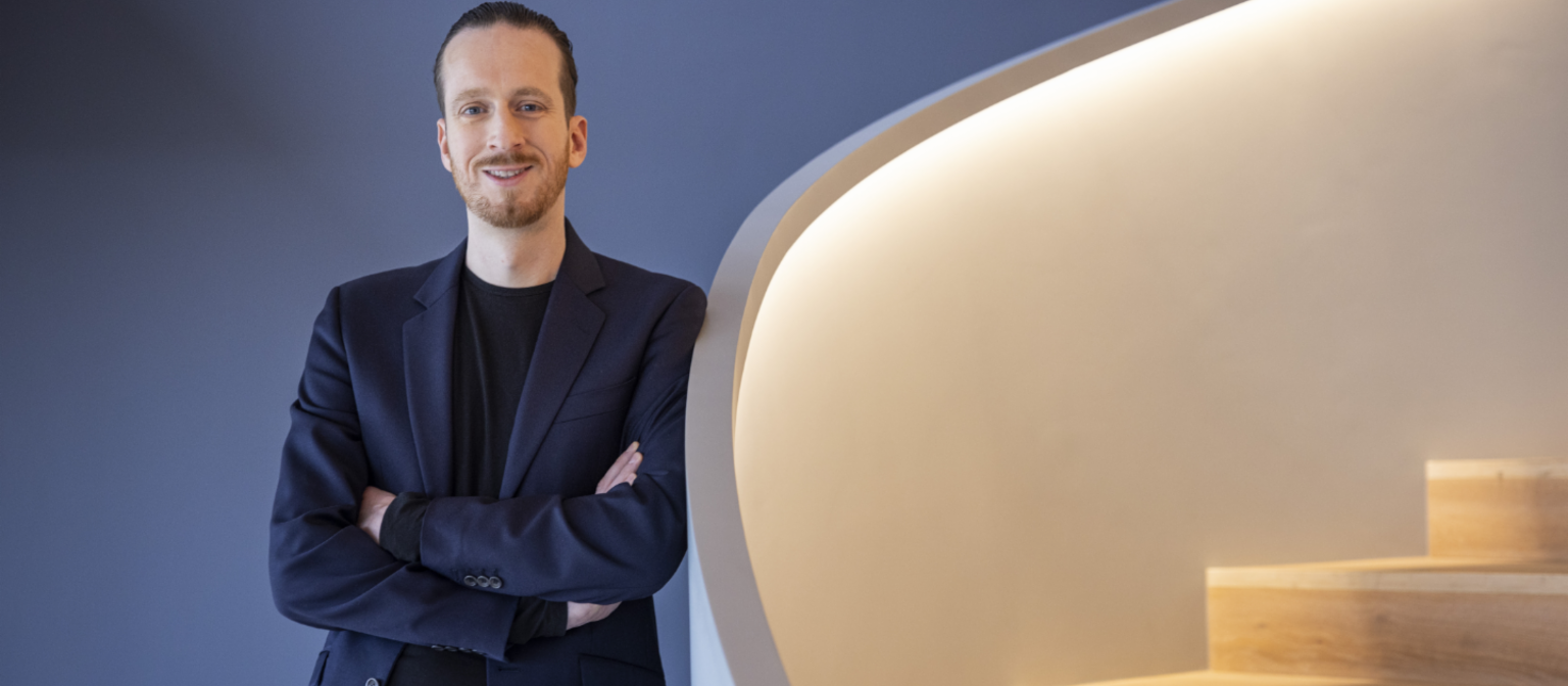 Sebastian Pollmer a male in a blue suit leans against a wall near to a white warm lightended stair