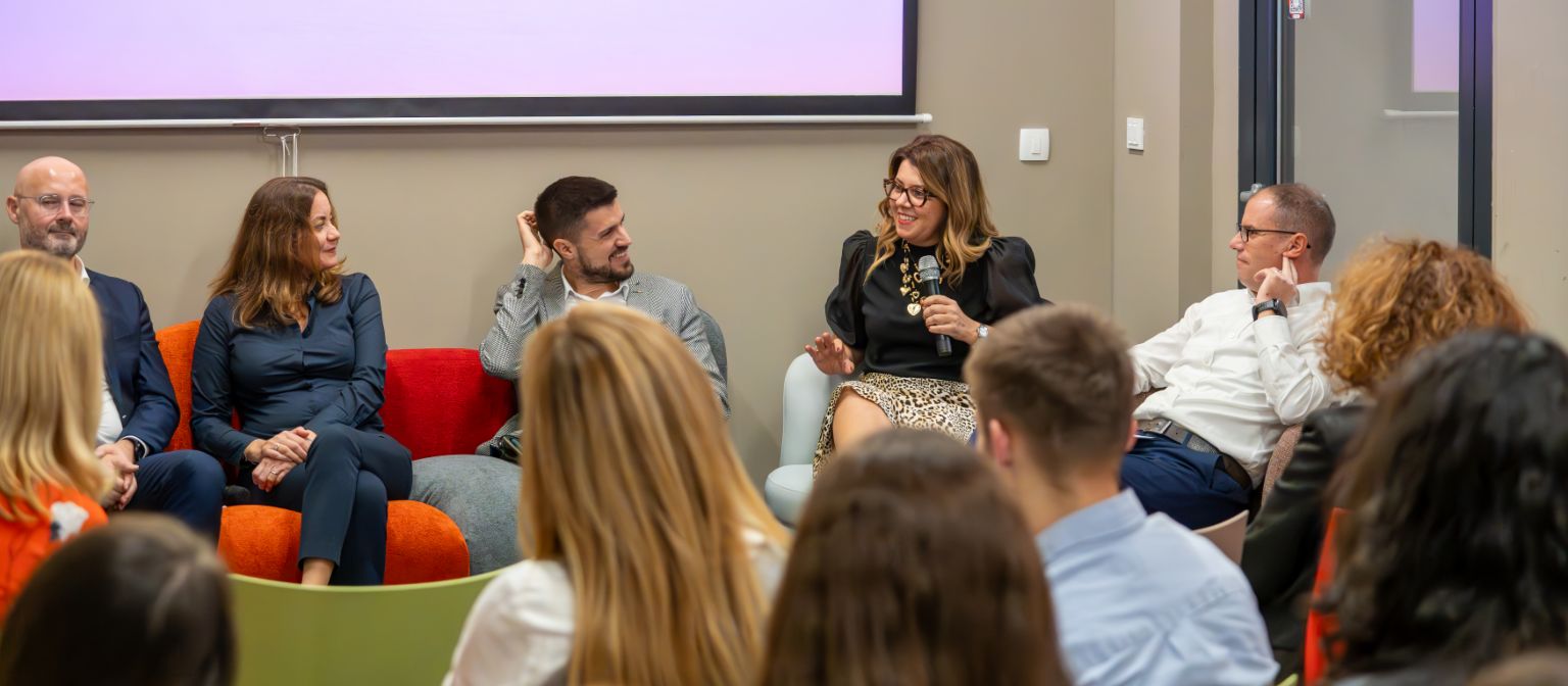 Panel diskusija s nekoliko govornika koji sjede na šarenoj sofi, a u pozadini je prikazana prezentacija. Fokus je na ženi koja govori, dok je publika uključena u raspravu.