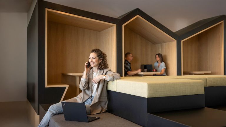 Nasmiješena osoba sjedi u modernom uredskom lounge prostoru, razgovara telefonom i radi na prijenosnom računalu. U pozadini rade još dvije osobe.