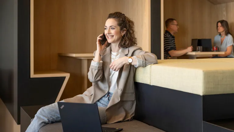 Nasmiješena osoba sjedi u modernom uredskom lounge prostoru, razgovara telefonom i radi na prijenosnom računalu. U pozadini rade još dvije osobe.