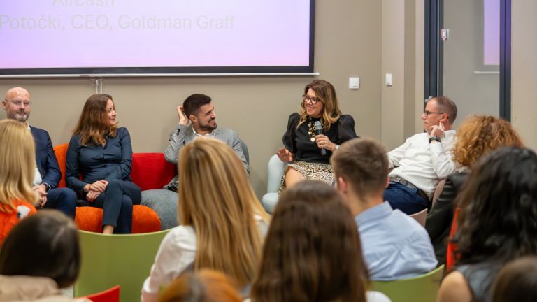 Panel diskusija s nekoliko govornika koji sjede na šarenoj sofi, a u pozadini je prikazana prezentacija. Fokus je na ženi koja govori, dok je publika uključena u raspravu.