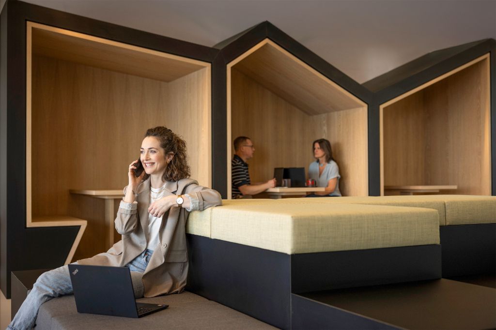 Nasmiješena osoba sjedi u modernom uredskom lounge prostoru, razgovara telefonom i radi na prijenosnom računalu. U pozadini rade još dvije osobe.